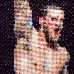 Champion — original 4×4 inch hand-stitched cross-stitch of a shirtless male athlete with a medal ribbon at his neck, stitched in warm peach, golden sand, blush, and sky blue on black Aida cloth